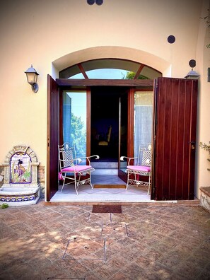 A building entrance with a wooden door, two white metal chairs with pink cushions, and a tiled patio area.