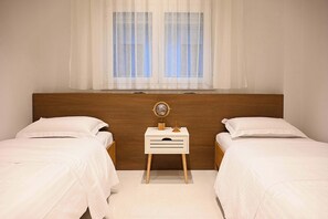 Ein Schlafzimmer mit zwei Einzelbetten, einem Holzkopfende, einem Nachttisch mit Globus und Lampe sowie einem Fenster mit transparenten Vorhängen.