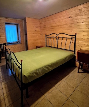 A single bed with a metal frame and a green bedspread in a room with wooden walls and tiled flooring. - Graziosa casa di montagna (Noasca)