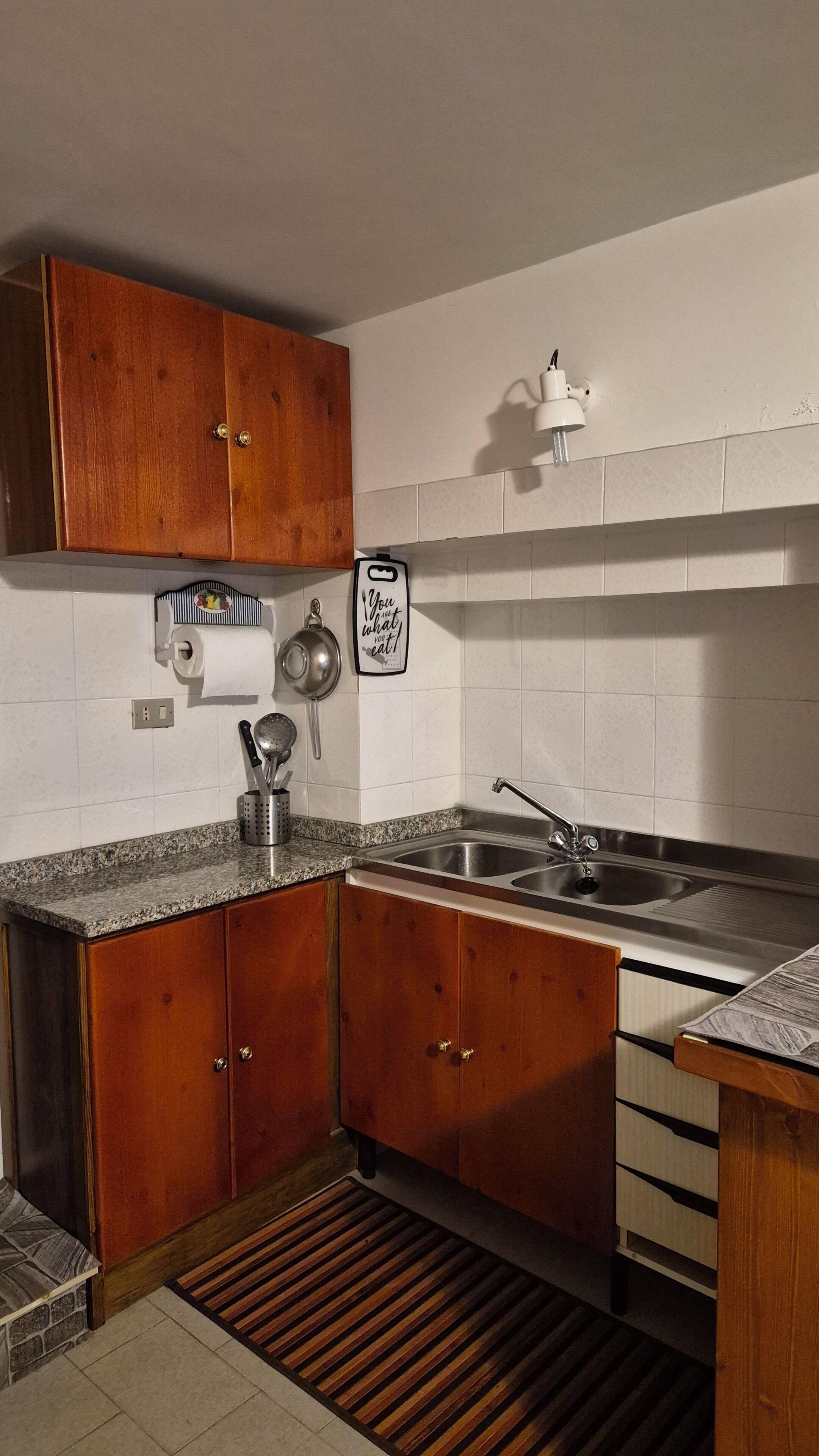 Cocina con gabinetes de madera, un lavamanos de acero inoxidable, una encimera con dispensador de toallas de papel y un accesorio de luz en la pared.