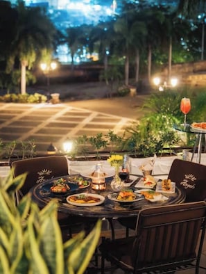 An outdoor dining area with tables set for a meal, surrounded by greenery and evening lighting. - Charming 2-bedroom Apartment in Santo Domingo Este (Santo Domingo Este)