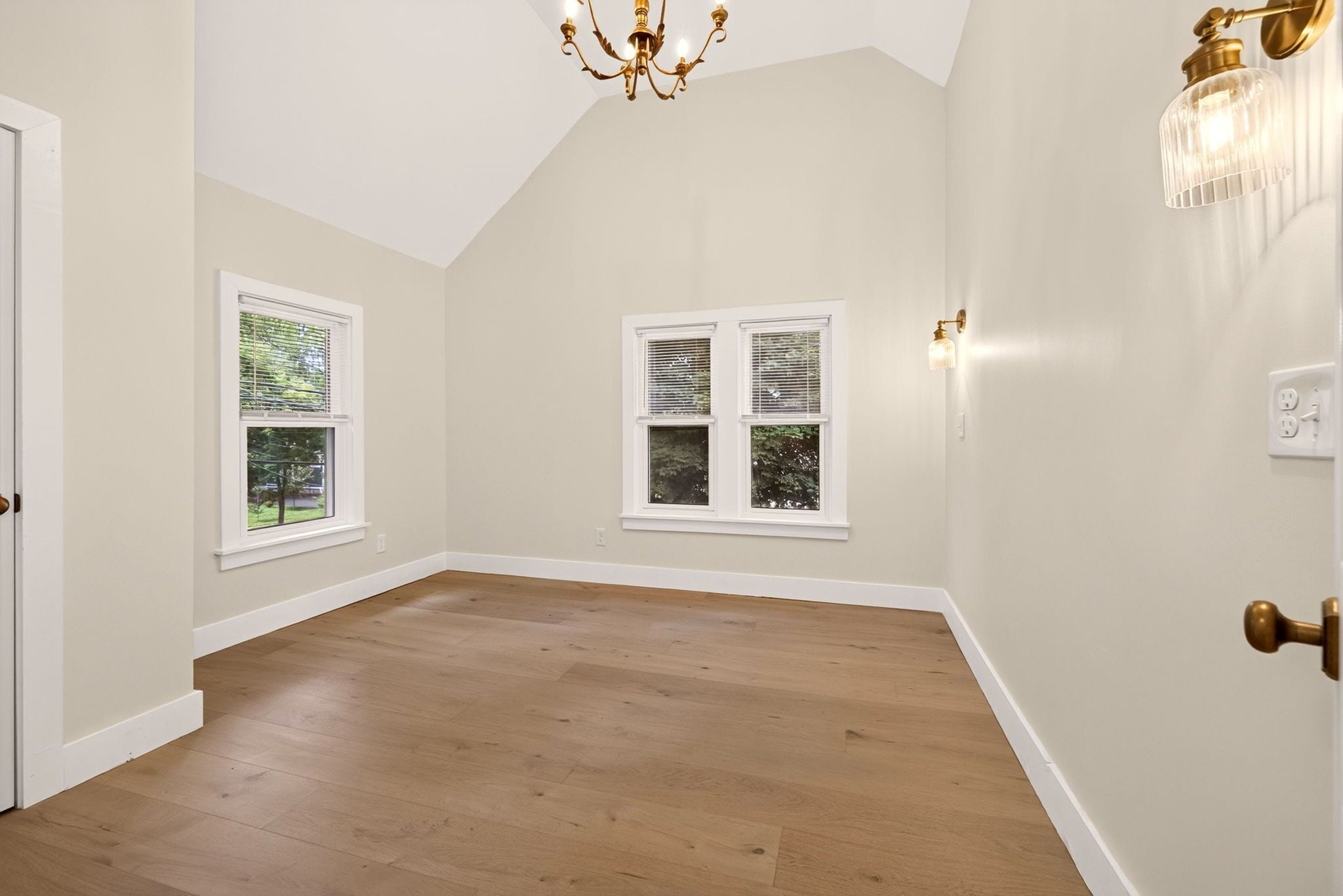 A room with wooden flooring, two windows with blinds, a chandelier, and a wall-mounted light fixture.