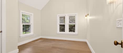 A room with wooden flooring, two windows with blinds, a chandelier, and a wall-mounted light fixture.