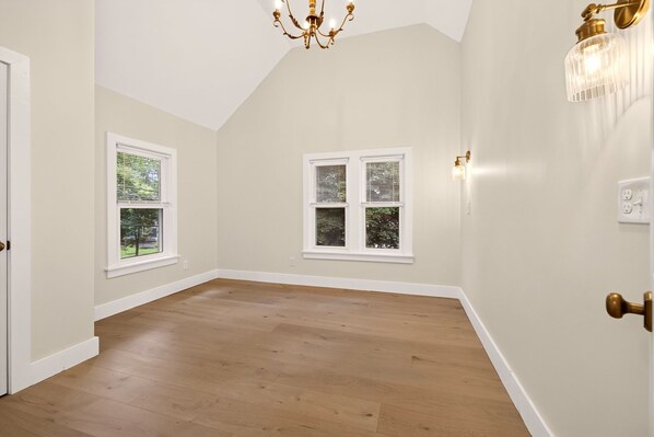 A room with wooden flooring, two windows with blinds, a chandelier, and a wall-mounted light fixture.