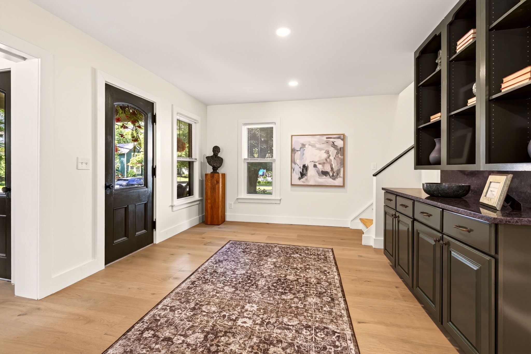 Un couloir avec un plancher en bois, un tapis à motifs, une porte sombre, un mur blanc avec une fenêtre et un meuble sombre sur lequel est posé un bol.