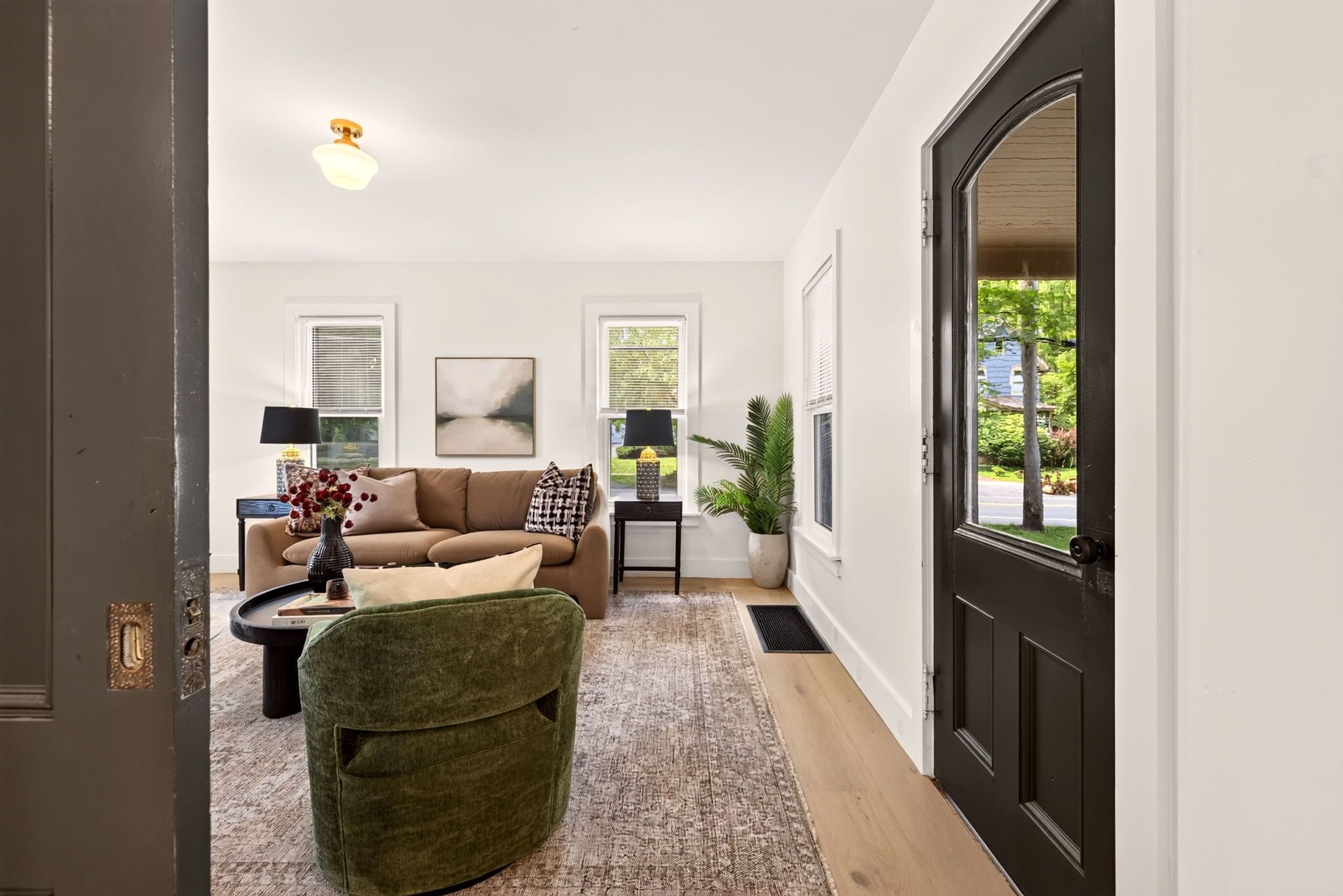 Un salon avec un canapé, un fauteuil vert, une table basse et une lampe. Il y a une porte sur la droite et une fenêtre sur la gauche.