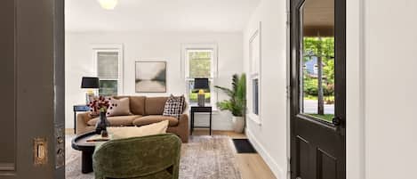 A living room with a sofa, a green armchair, a coffee table, and a lamp. There is a door on the right and a window on the left.