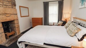 Ein Schlafzimmer mit Kamin aus Stein, einem Bett mit weißer Bettwäsche, einer hölzernen Kommode und einem Sessel mit gemusterten Kissen.