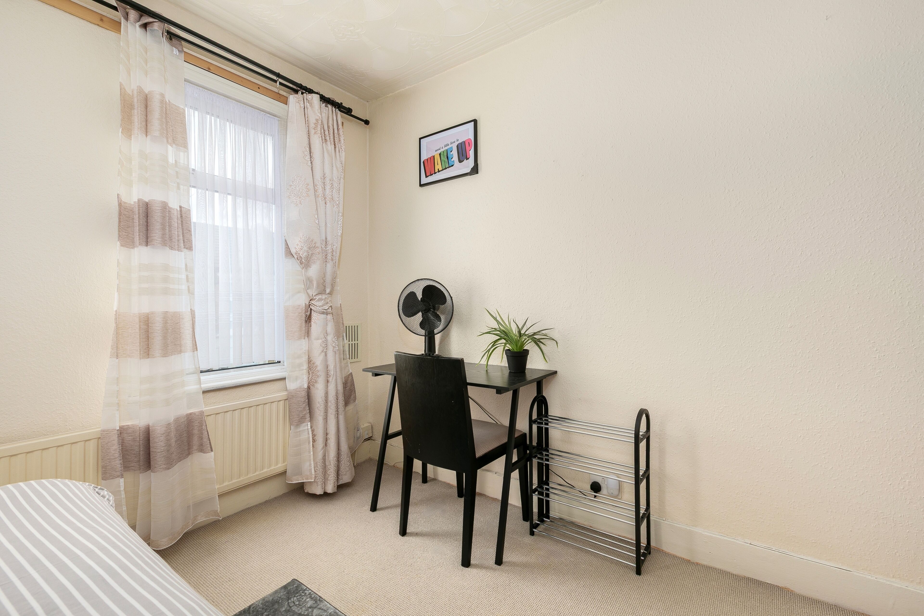 A room with a bed, a desk, a chair, a radiator, a fan, a potted plant, and a picture on the wall.