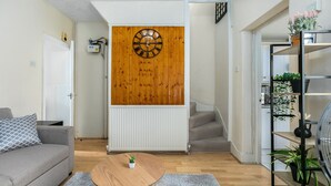 Ein Wohnzimmer mit einer Holz-Wanduhren, einem grauen Sofa, einem runden Holztisch und einer Treppe, die in einen anderen Raum führt.