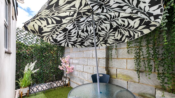 Ein runder Glastisch mit einem Stuhl aus Metall, ein schwarz-weiß gemusterter Schirm und eine kleine, blühende rosa Pflanze.