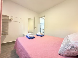 Quarto com colcha rosa, espelho, guarda-roupa e porta que dá para outro cômodo.