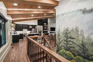 A modern kitchen with dark cabinetry, a wooden ceiling, and a dining area with a view of a forest mural. - The Rock Haus | Pool, Putt-Putt | Hocking Hills (Laurelville)