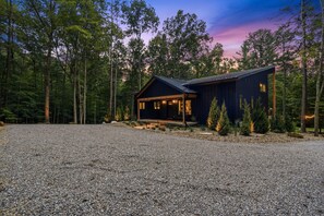 A cabin with a gravel driveway, surrounded by tall trees and a clear sky. - The Rock Haus | Pool, Putt-Putt | Hocking Hills (Laurelville)