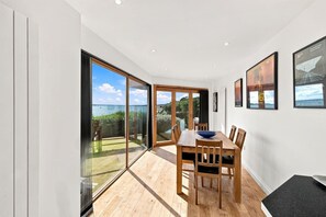 Uma sala de jantar com mesa e cadeiras de madeira, grandes portas de vidro deslizantes que dão para um jardim, e quadros nas paredes.