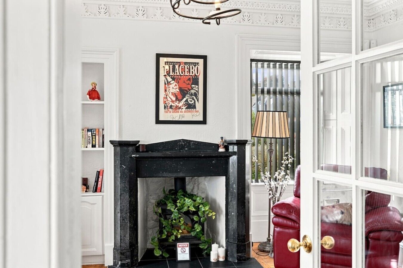 Een kamer met een open haard, een ingelijste poster aan de wand, een boekenplank en een rode fauteuil.
