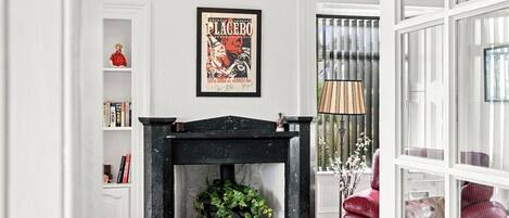 Een kamer met een open haard, een ingelijste poster aan de wand, een boekenplank en een rode fauteuil.