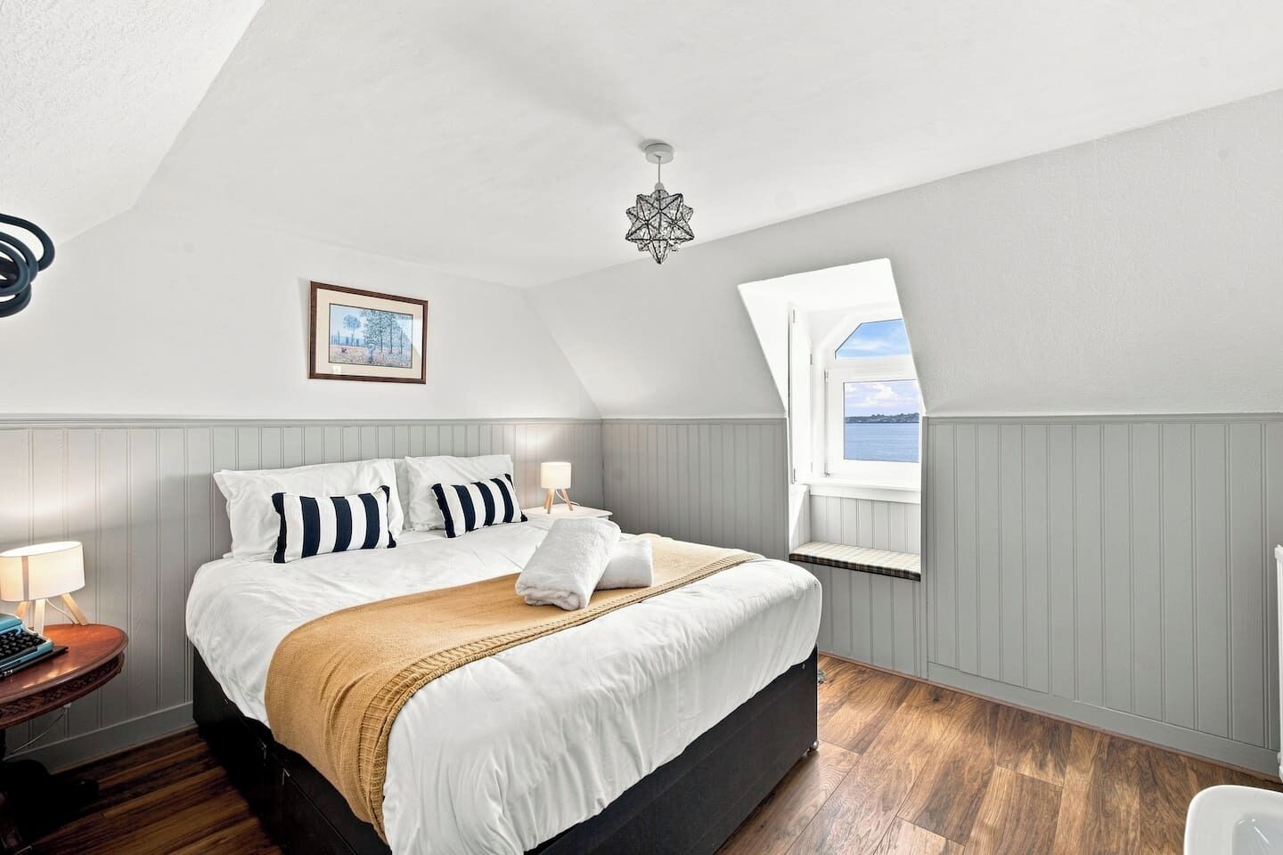 A bedroom with a bed, a bedside table, a lamp, a framed picture, a window with a view, and a star-shaped light fixture.