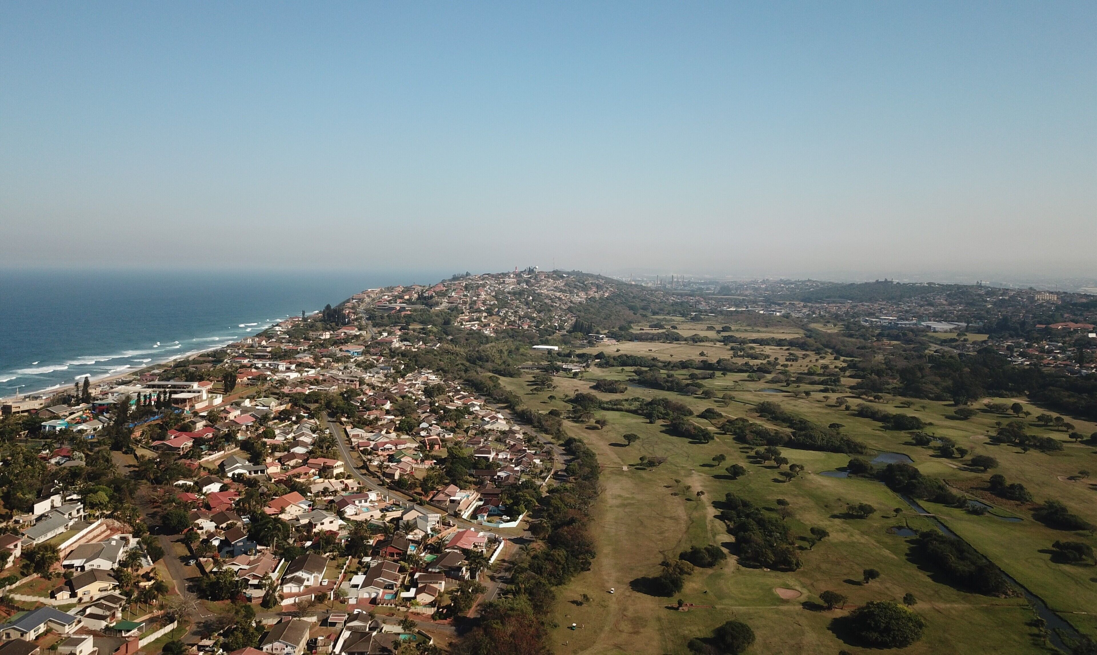 Vista aèria d'una ciutat costanera amb zones residencials, un camp de golf i un paisatge amb turons.
