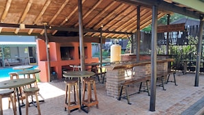 Un patio al aire libre con piso de ladrillo, mesas y taburetes de madera, una parrilla de ladrillo y una piscina al fondo.