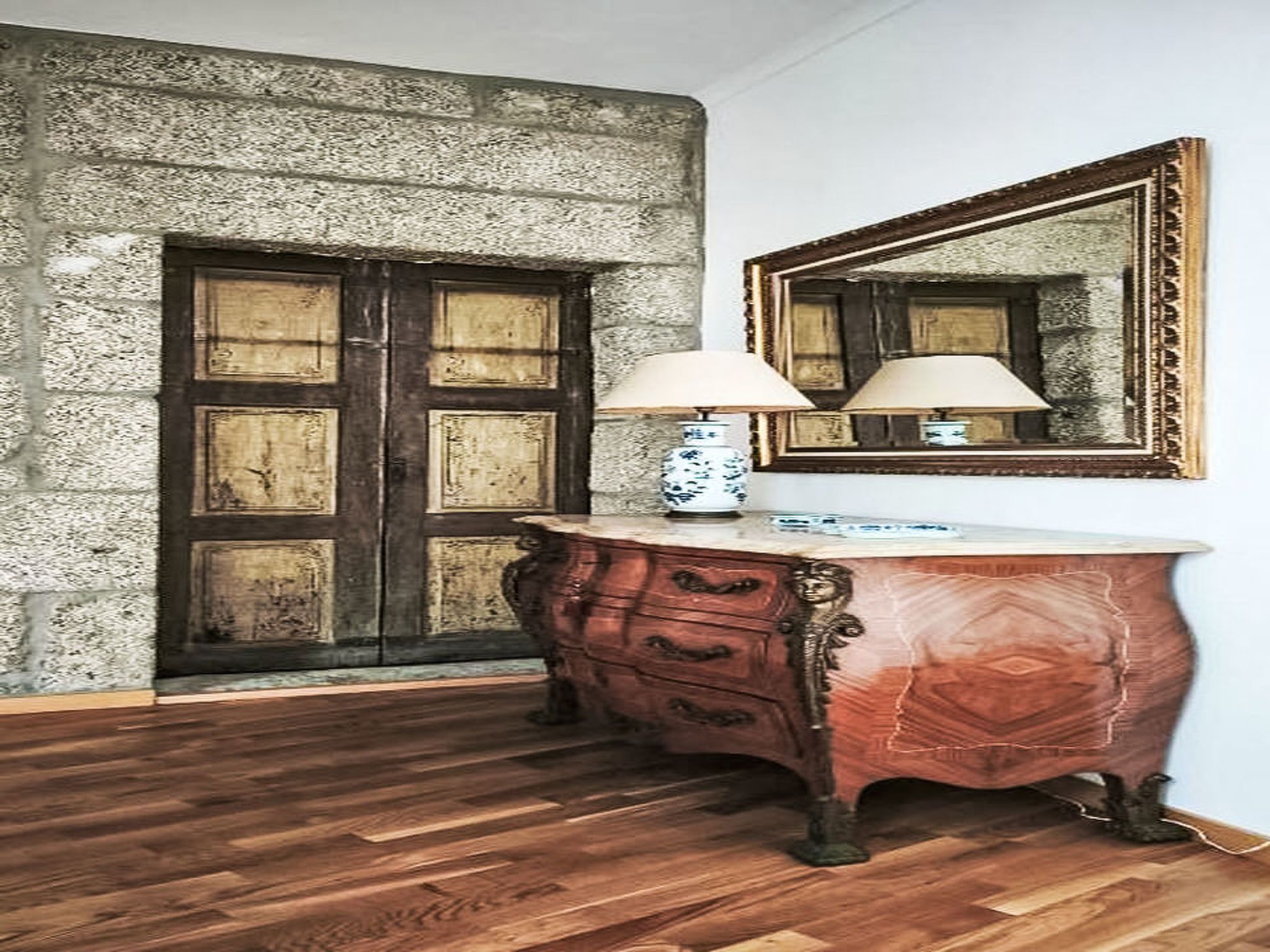 Una stanza con pavimento in legno, parete in pietra, porta in legno, un comò con cassetti e uno specchio.