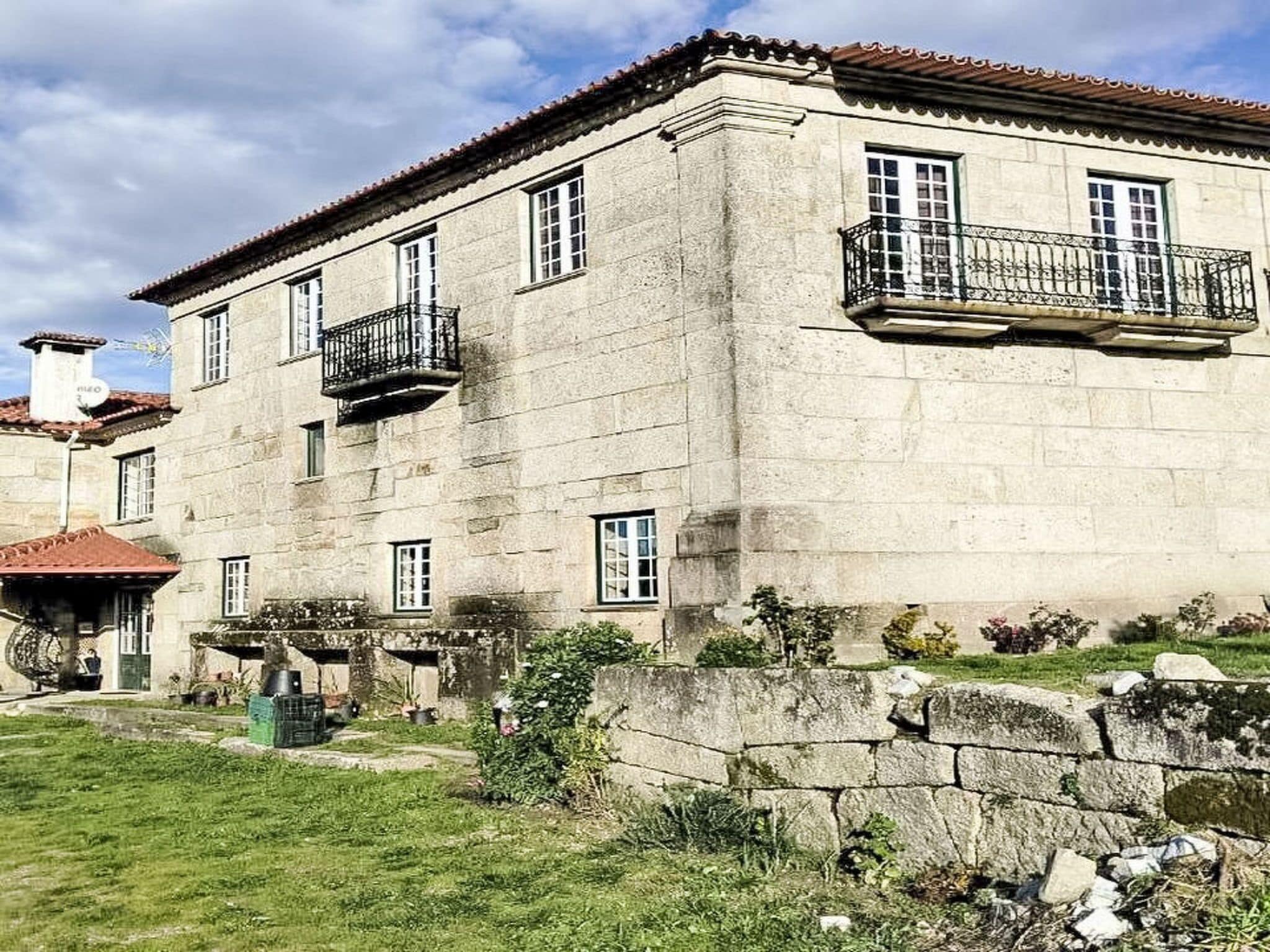 Un bâtiment en pierre de deux étages, avec un toit recouvert de tuiles rouges, un mur en pierre et un petit jardin.