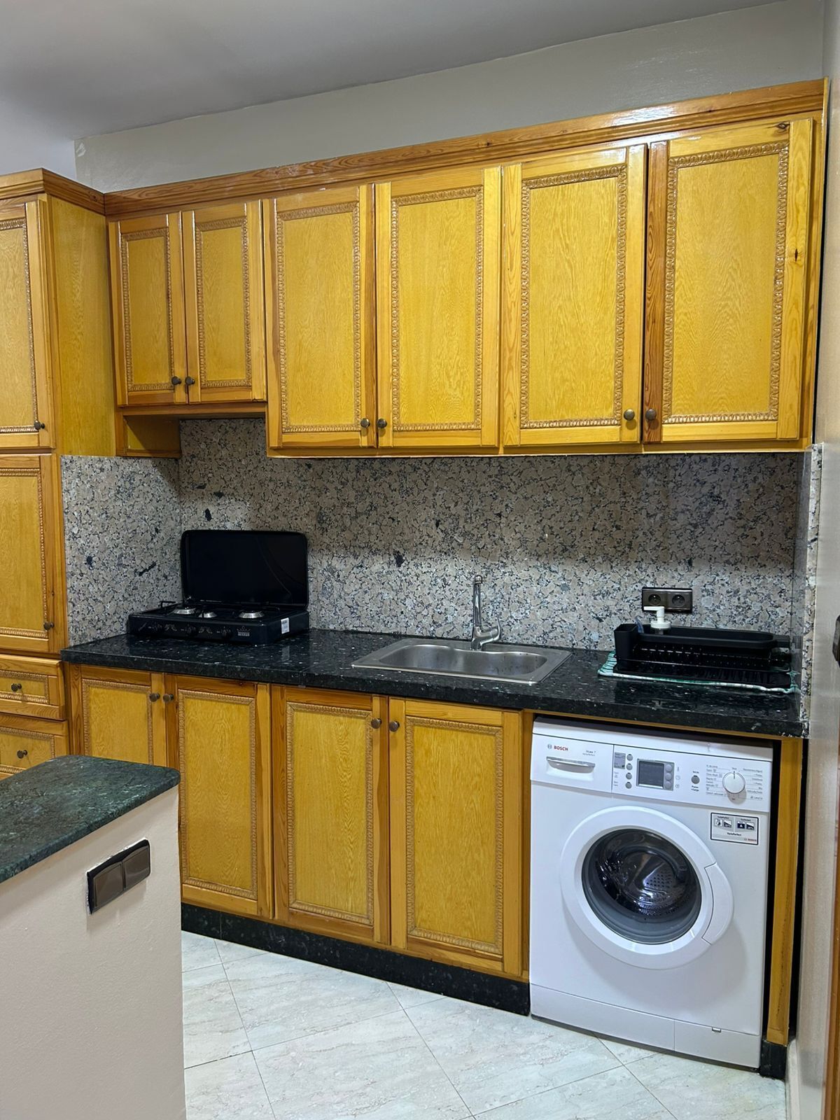 Una cocina con armarios de madera, encimera negra, pileta de acero inoxidable y lavadora Bosch blanca.
