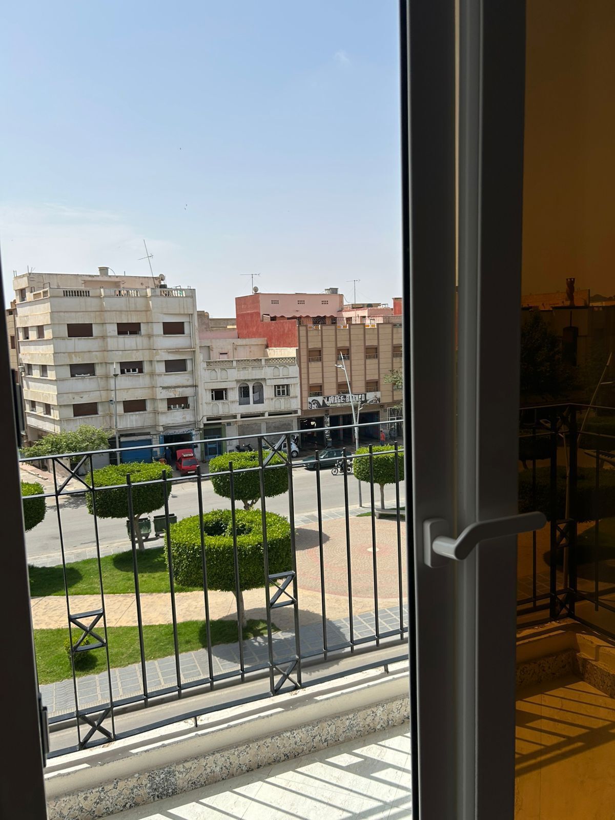 Une vue depuis un balcon doté d’une rambarde en métal, donnant sur une rue bordée d’immeubles et un ciel dégagé.