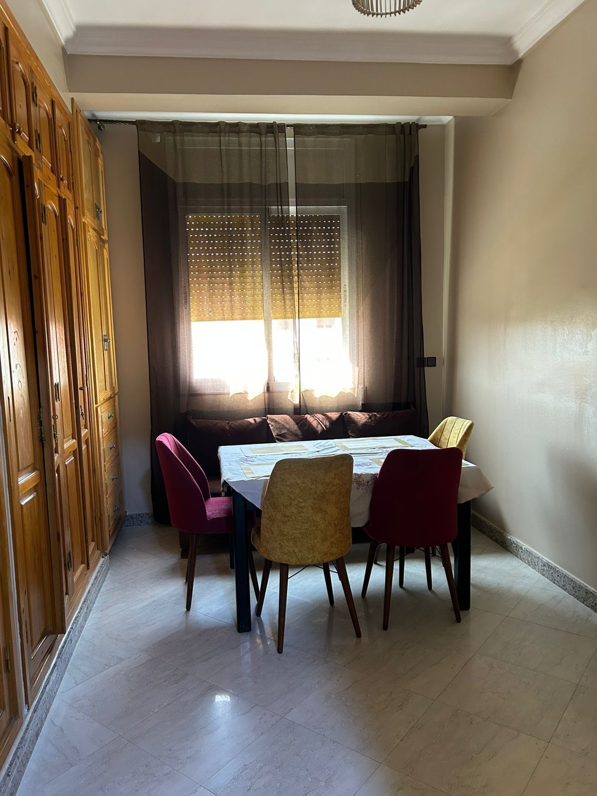A dining area with a table set for four, a window with blinds, and a wooden cabinet to the left.