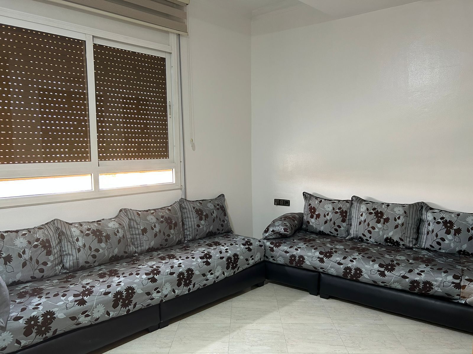A corner room with a sectional sofa, a window with blinds, and a wall-mounted thermostat.