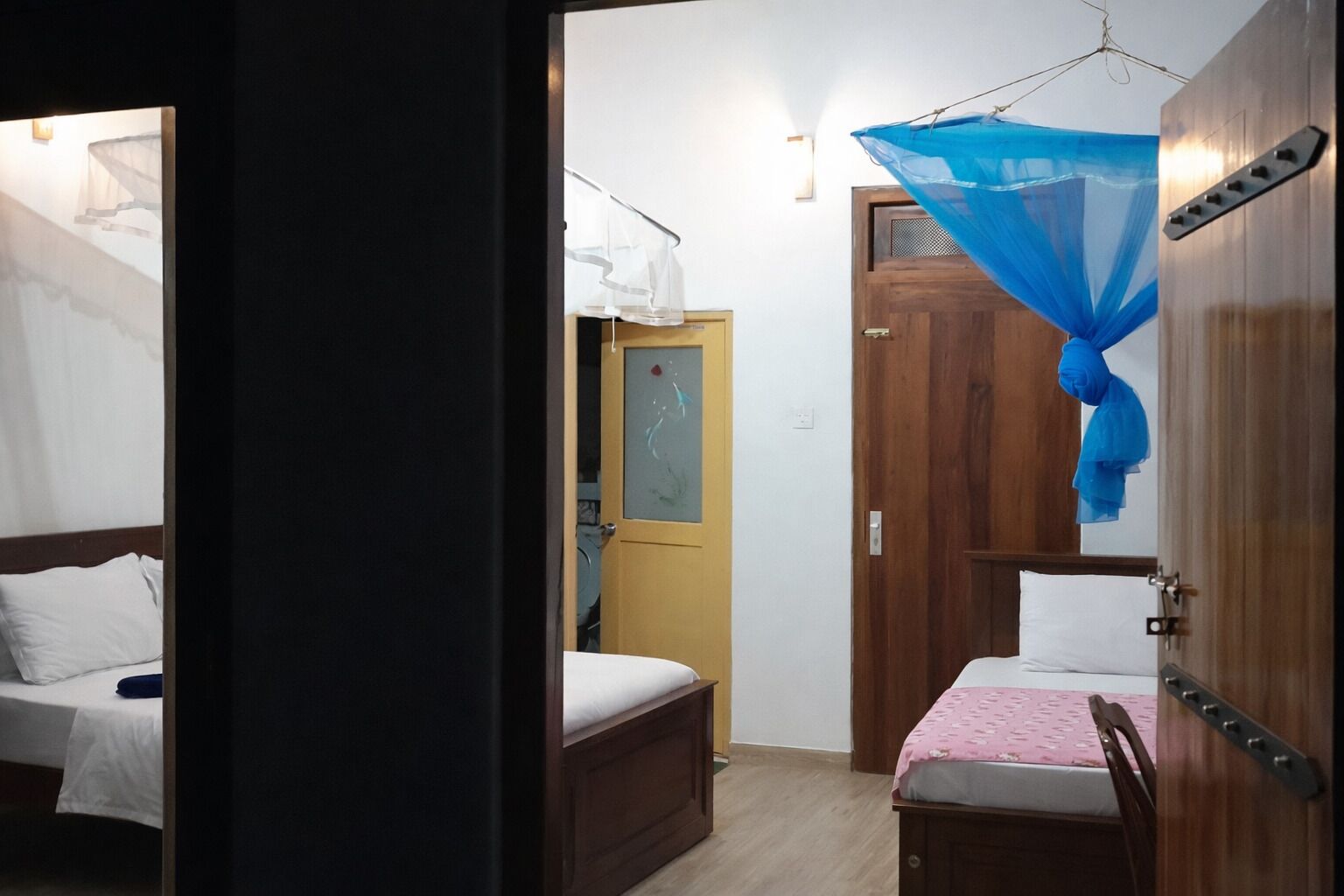 A hotel room with two beds, a wooden wardrobe, a door with a glass panel, and a blue mosquito net.