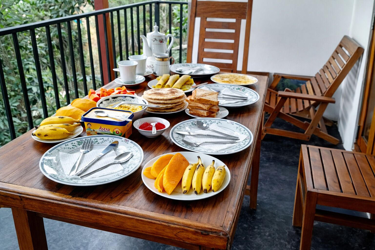 Une table en bois, dressée avec des assiettes, des couverts et des aliments comme des bananes et du pain, sur un balcon doté d’une balustrade et de chaises.