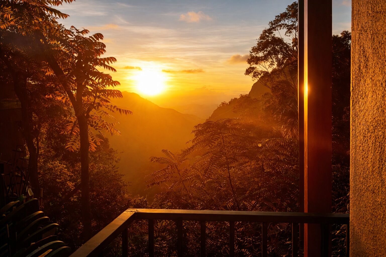 Solnedgangsudsigten fra en balkon med gelænder, over et dalområde med frodig vegetation og et fjerntliggende bjerg.