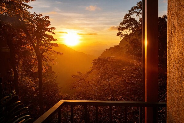 A sunset view from a balcony with a railing, overlooking a valley with dense foliage and a mountain in the distance.