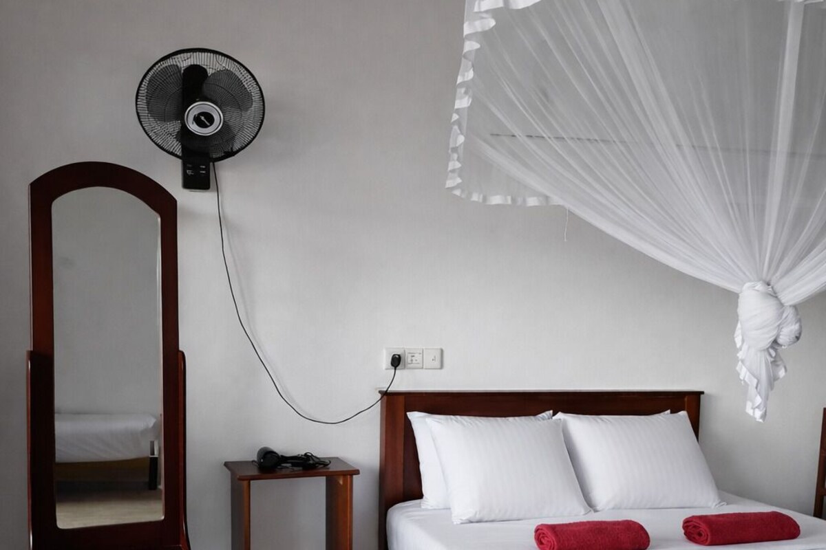 A bedroom with a bed, a bedside table, a mirror, a standing fan, and a mosquito net.