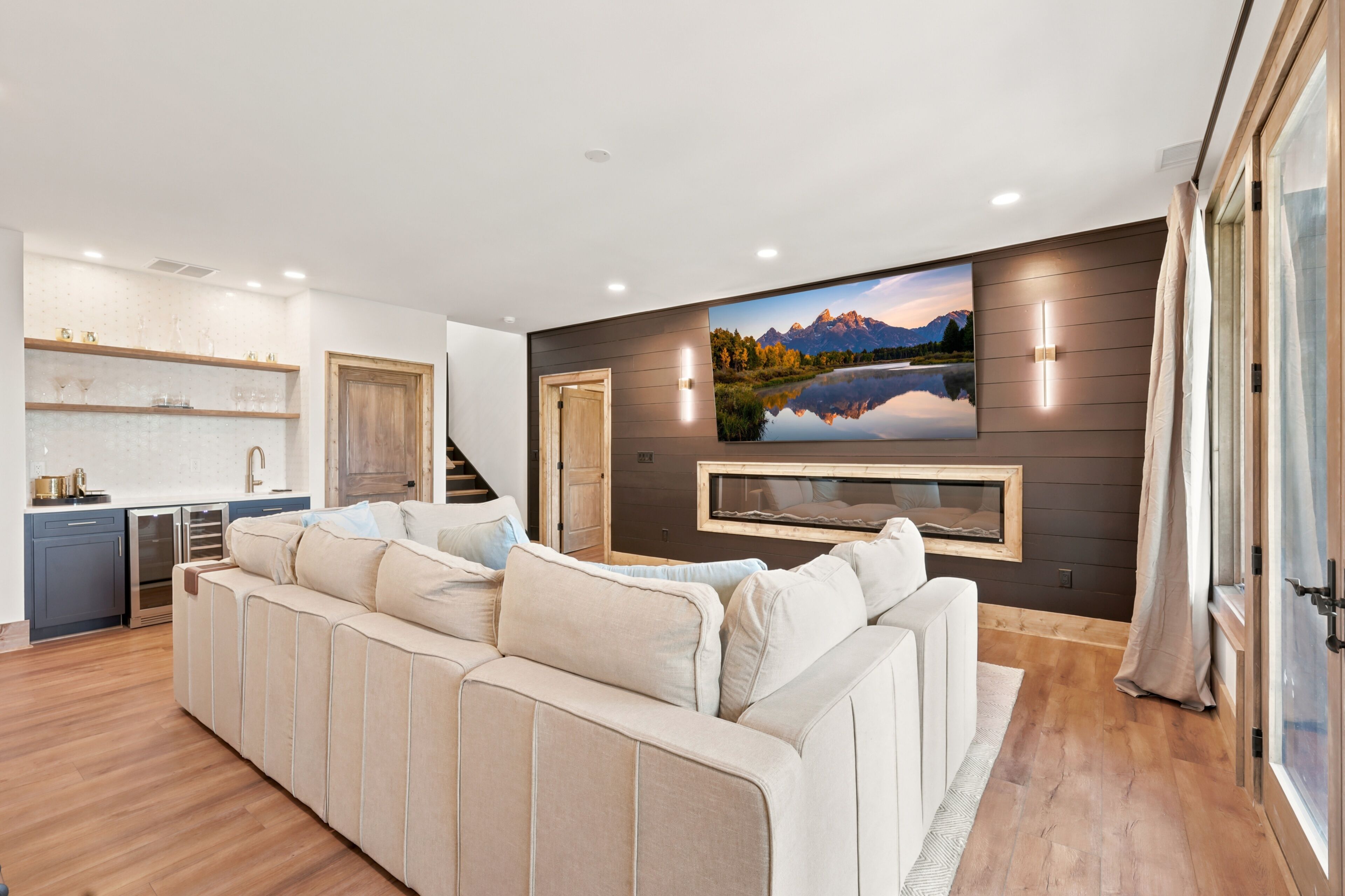 Ein modernes Wohnzimmer mit einem großen Ecksofa, einer Küchenzeile mit Einbaugeräten und einem großen, an der Wand befestigten Fernseher, der eine schöne Landschaft zeigt.