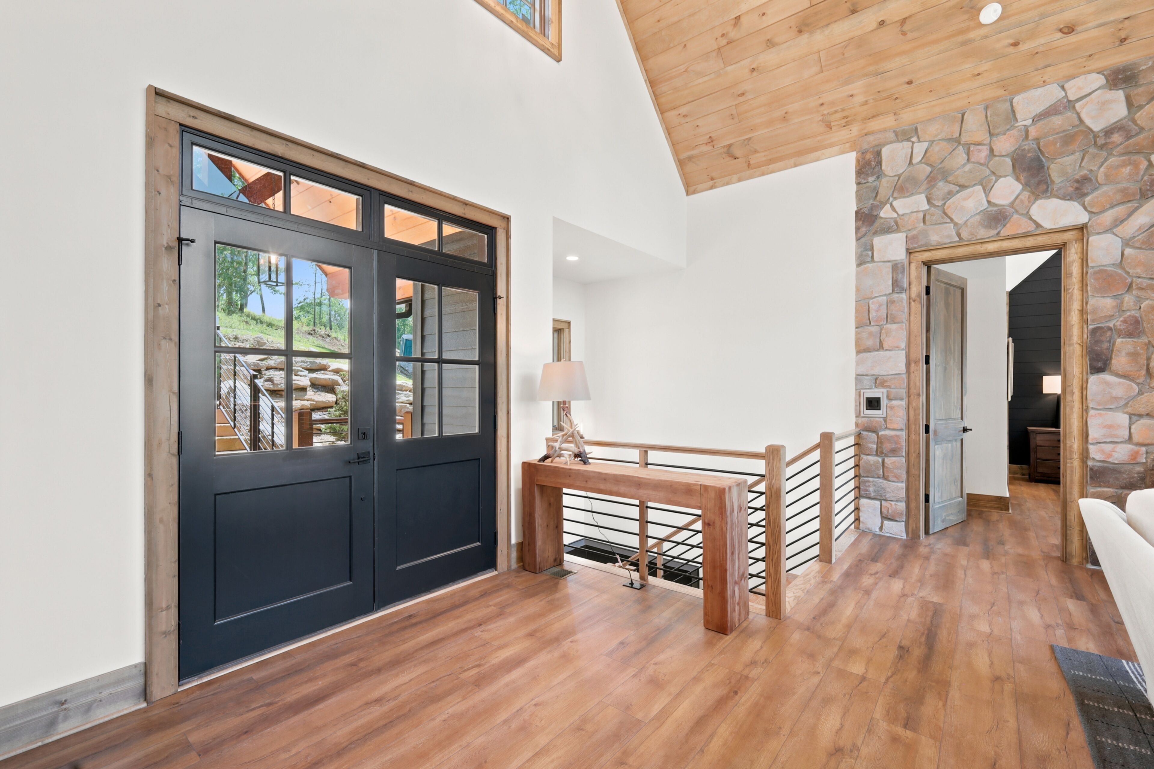A room with a large black door, wooden flooring, a stone wall, and a staircase leading to another level.