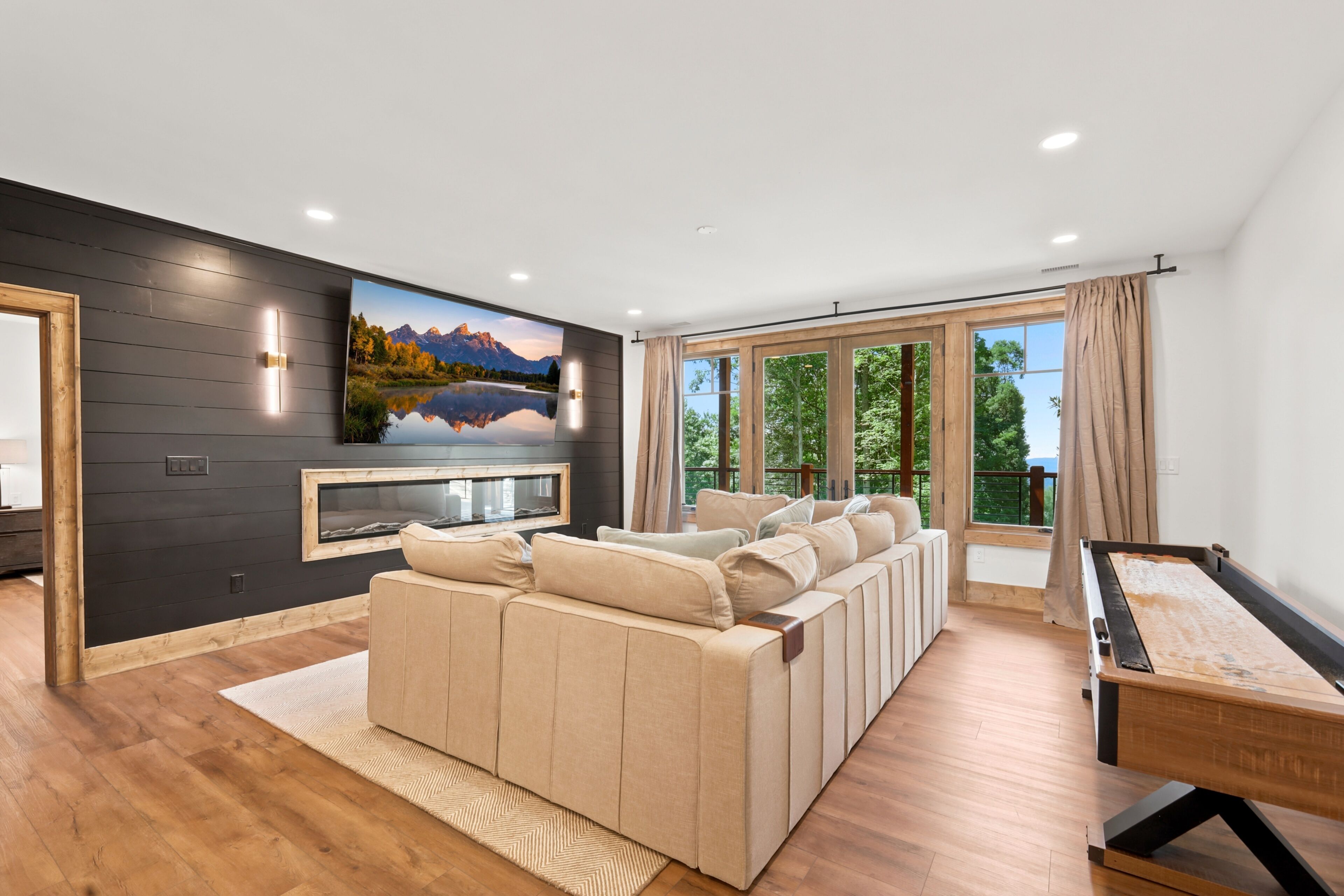 A modern living room with a large sectional sofa, a flat-screen TV mounted on a dark wall, and a pool table.