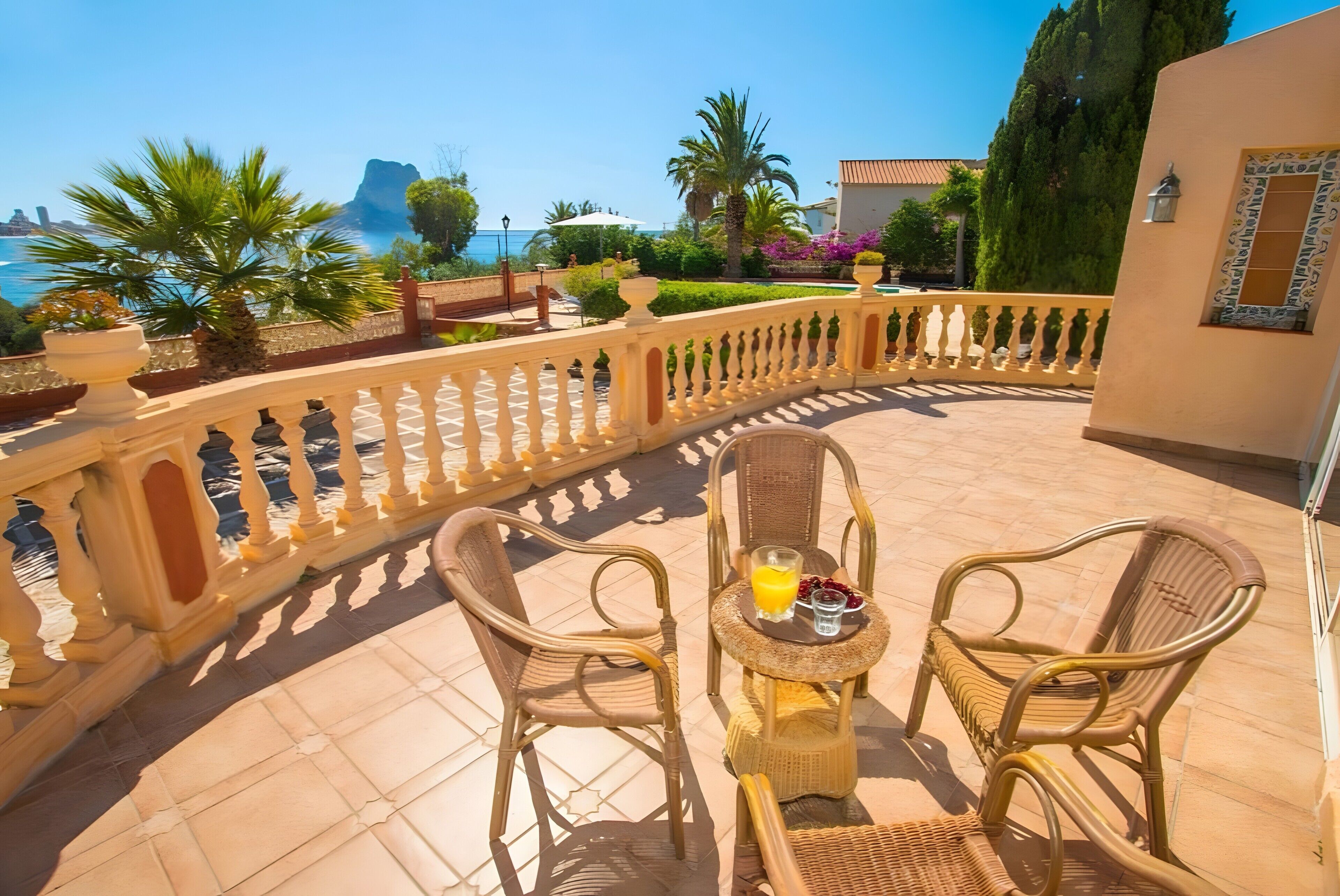 Un área de estar al aire libre con forma curva, con dos sillones de mimbre y una mesa, con vistas a un paisaje costero con una montaña al fondo.