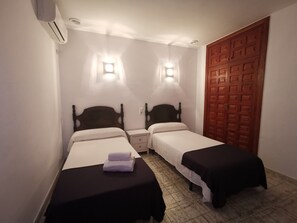 A hotel room with two single beds, a nightstand, a wooden door, and a wall-mounted air conditioning unit.