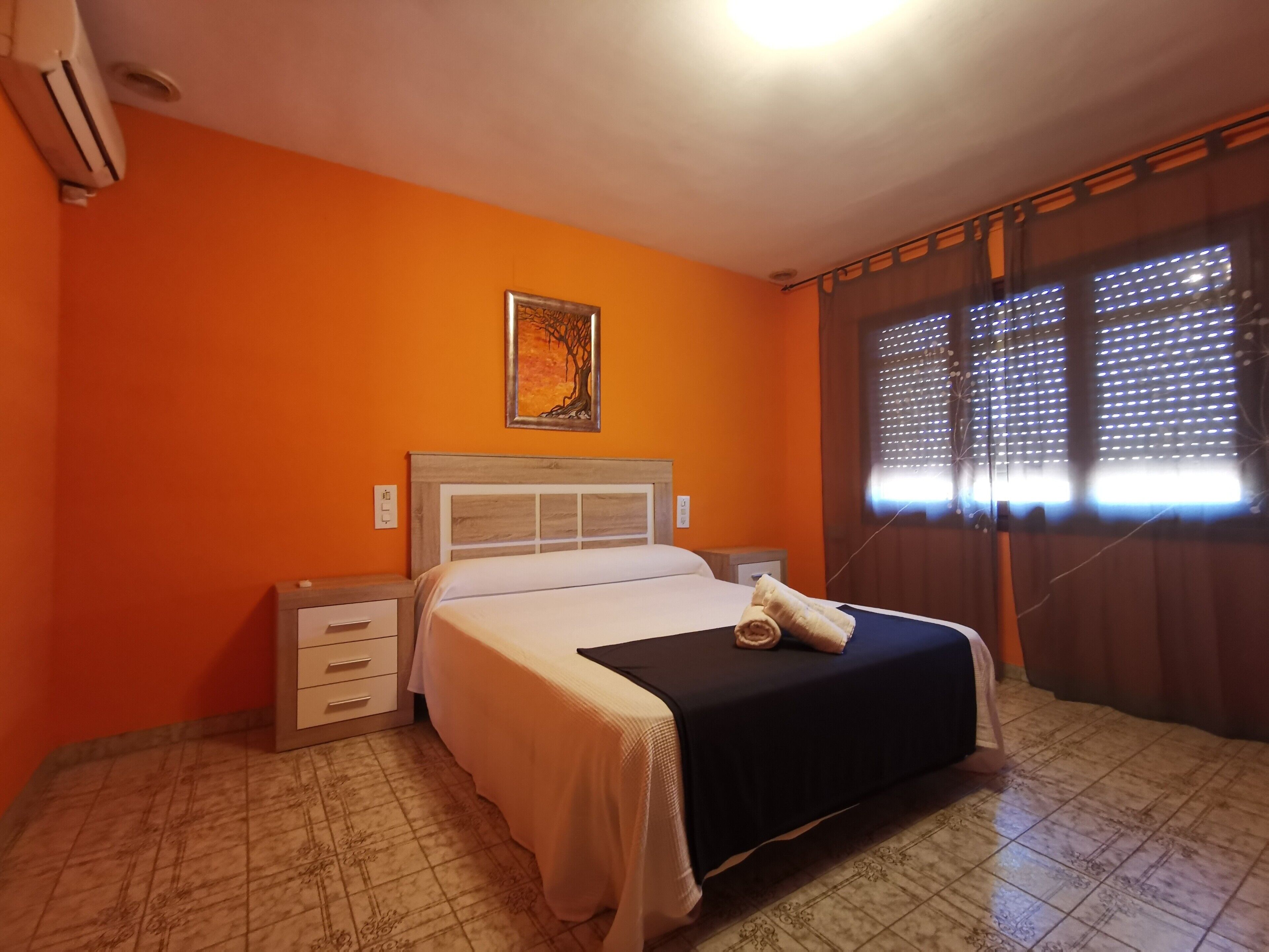 A hotel room with a bed, a nightstand, a drawer, a painting on the wall, and a window with blinds.