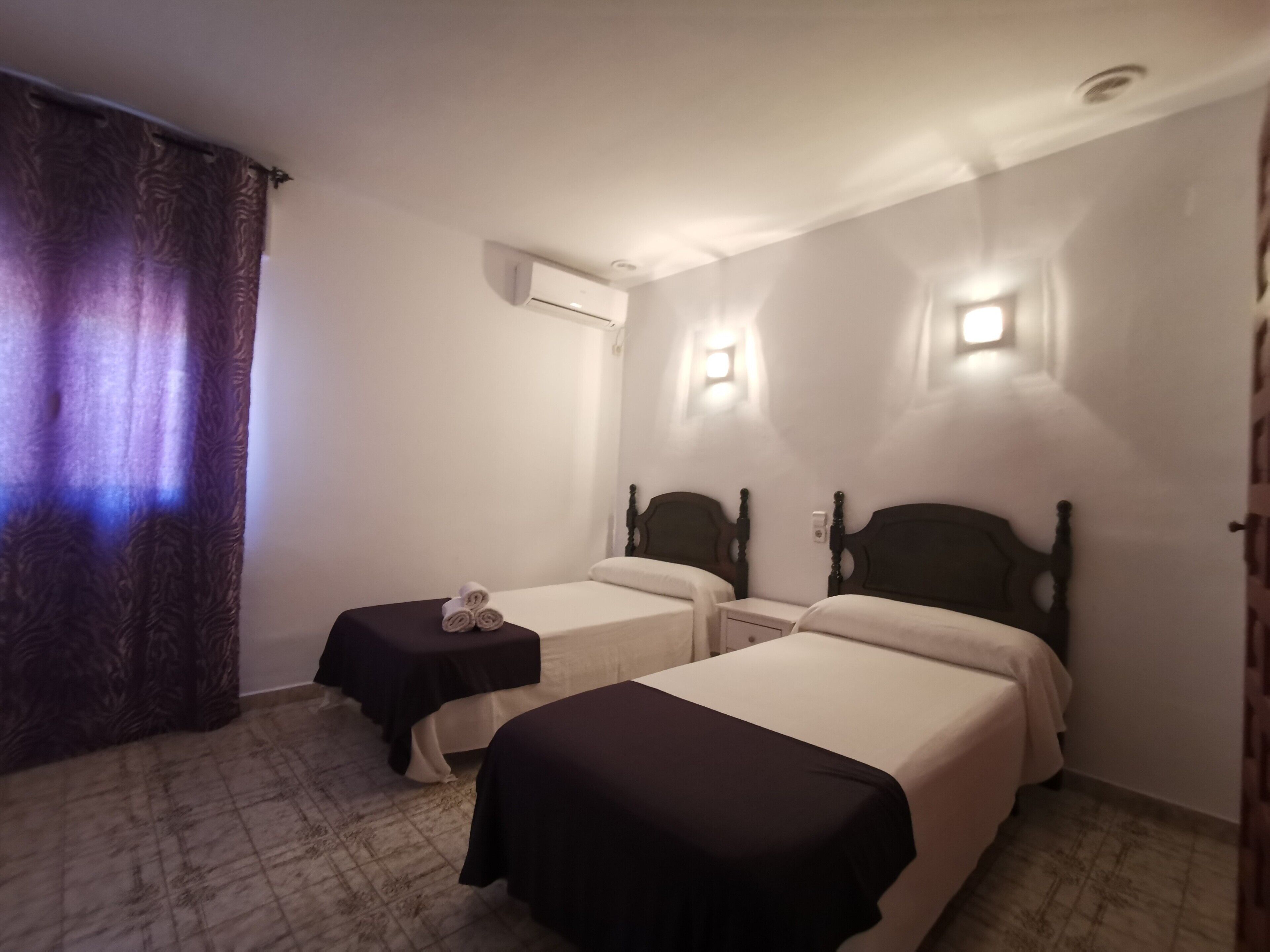 A hotel room with two single beds, a small table with a towel, an air conditioning unit, and two wall-mounted lights.