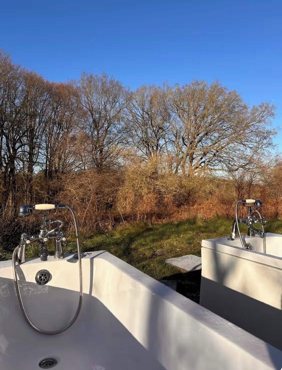Deux éviers extérieurs, équipés de robinets et de tuyaux, sont entourés d’arbres et d’arbustes.