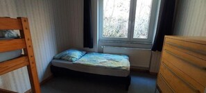 A small room with a single bed, a wooden dresser, and a window with a view of trees.