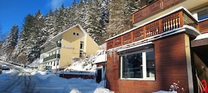 A building with a wooden facade and a large window, surrounded by snow-covered trees and a clear sky.