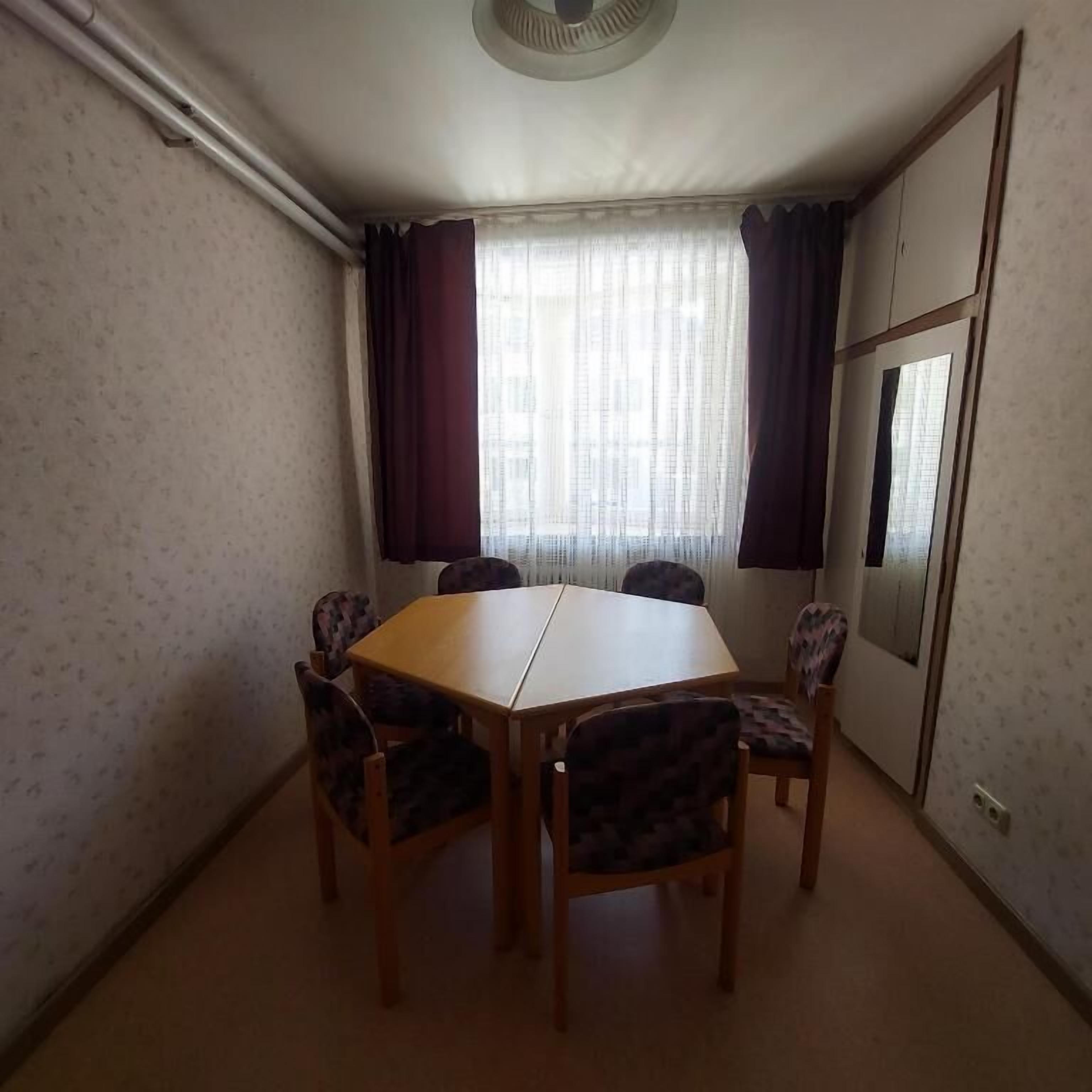 Ein Zimmer mit einem Holztisch, vier Stühlen, einem mit Vorhängen versehenen Fenster und einer Spiegelzierde an der Tür.