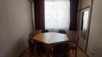 Ein Zimmer mit einem Holztisch, vier Stühlen, einem mit Vorhängen versehenen Fenster und einer Spiegelzierde an der Tür.
