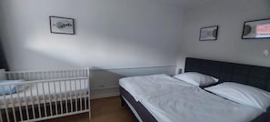 A bedroom with a white crib, a bed with white bedding, and two framed pictures on the wall. - Familienapartments Wildemann (Wildemann)