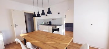 A modern kitchen with a wooden dining table, white chairs, and pendant lighting.