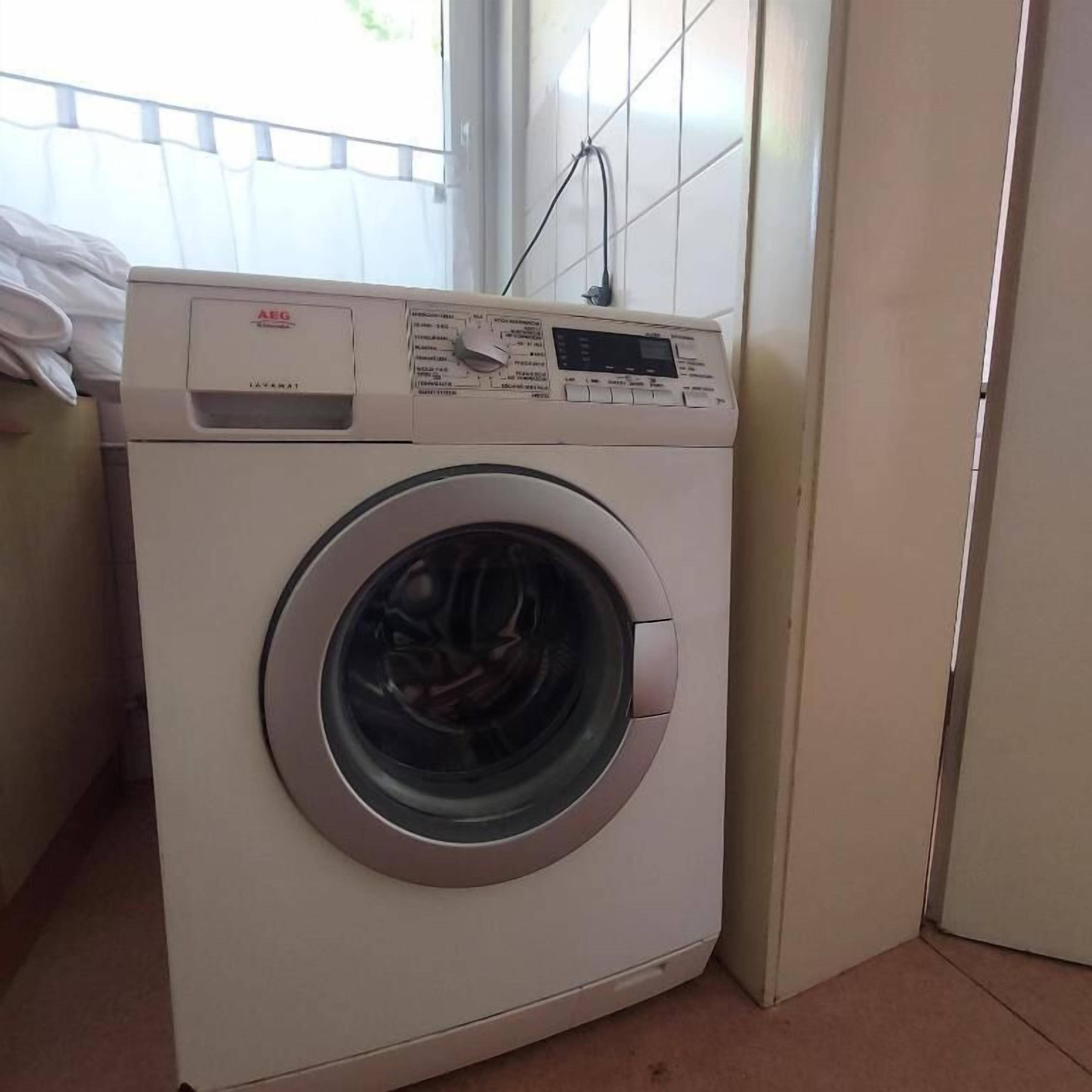 Eine Waschmaschine mit Frontlader und Bedienfeld, platziert in einer Ecke eines Raumes mit gefliesten Wänden und einem Fenster.